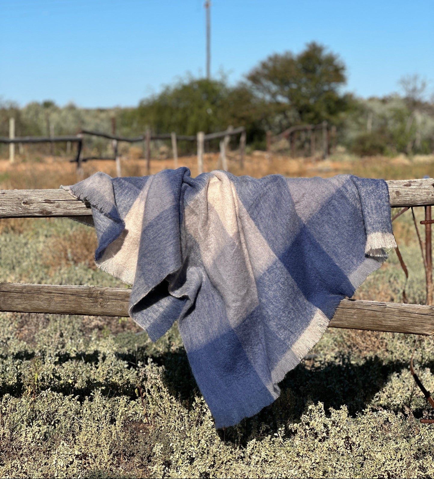 Blue and gray striped blanket draped over a wooden fence in a rural setting with trees and clear sky.
