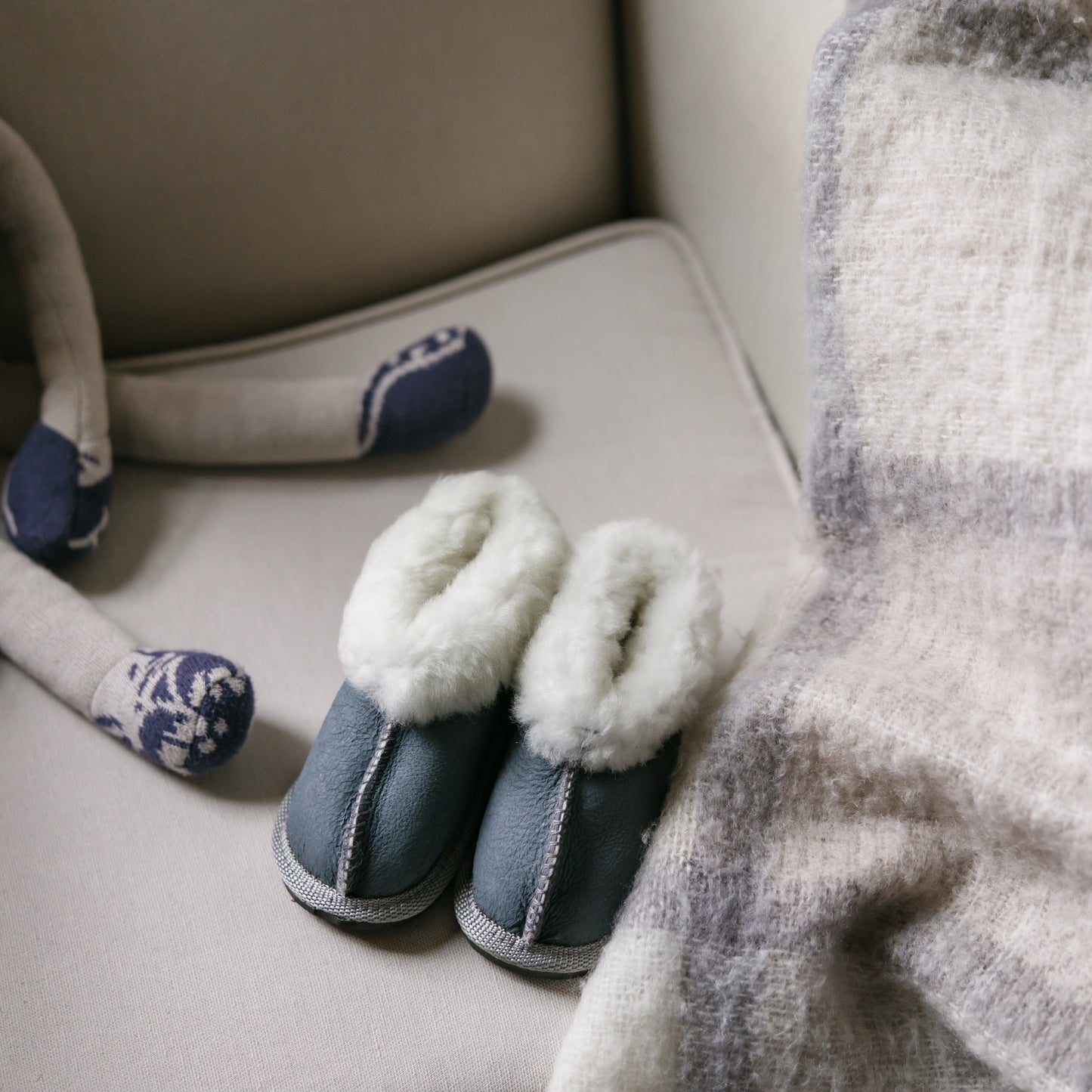 Pair of blue slippers on a beige surface with a plaid blanket and stuffed animal in the background.