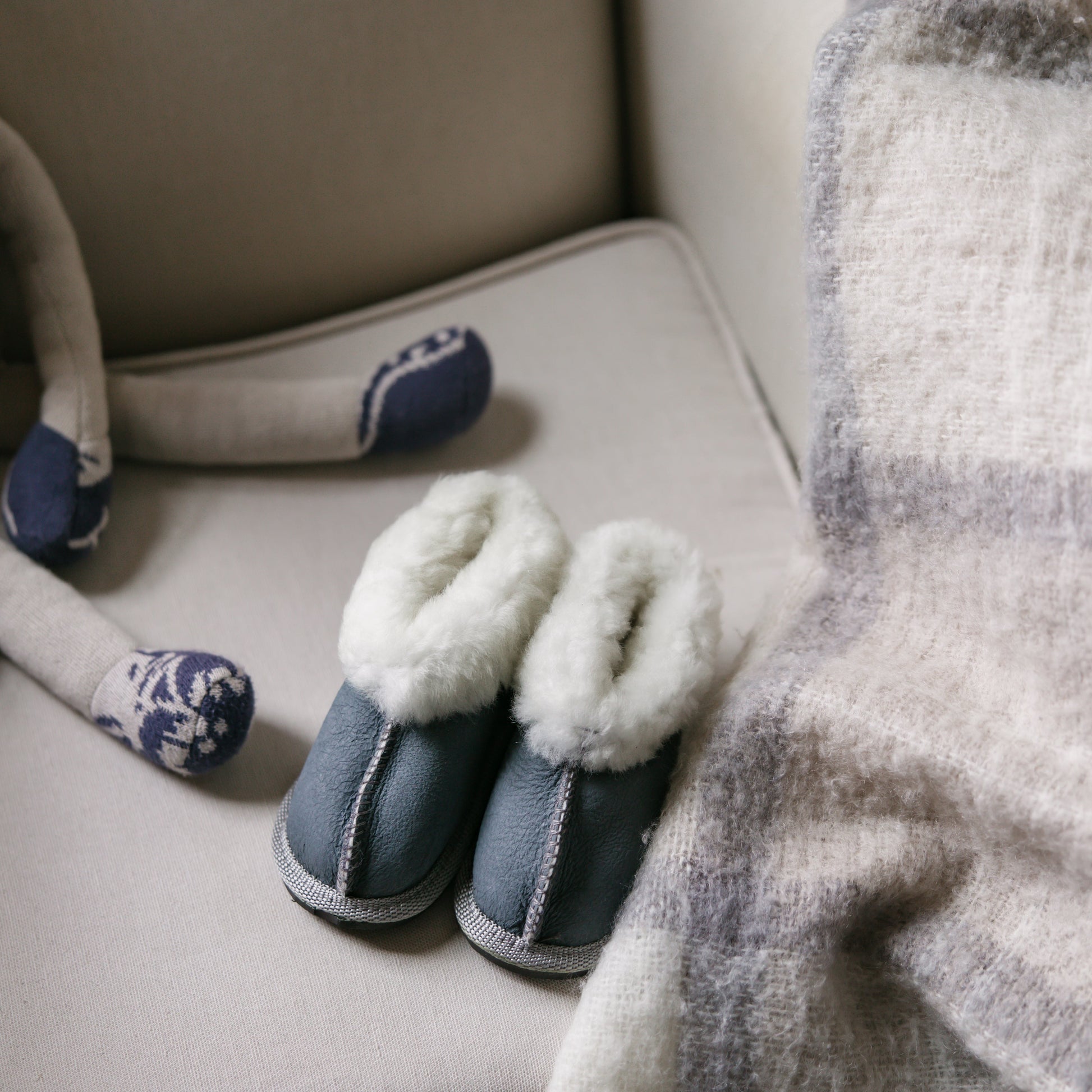 Pair of blue slippers on a beige surface with a plaid blanket and stuffed animal in the background.