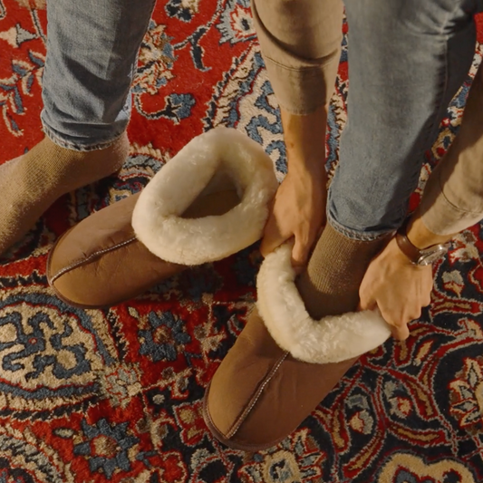 Person putting on brown slippers with white fur trim on a patterned rug