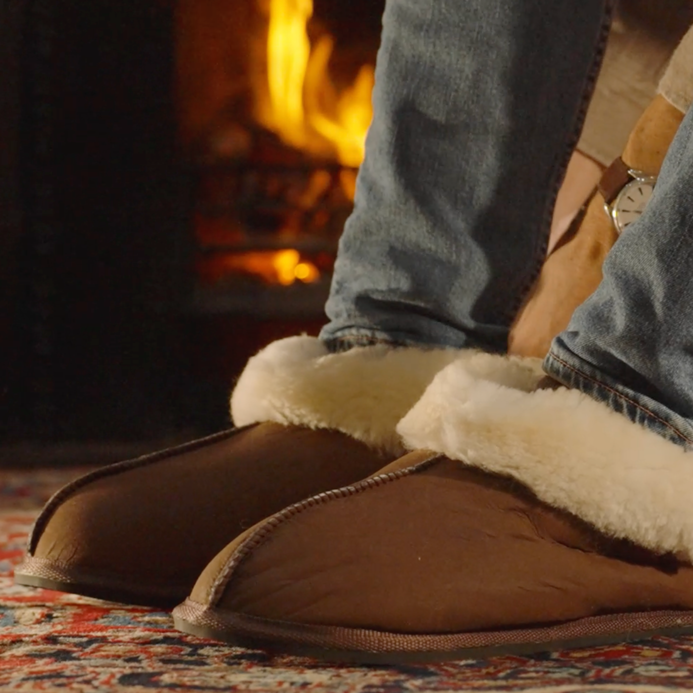 Brown slippers with white fur trim worn by a person in front of a fireplace.