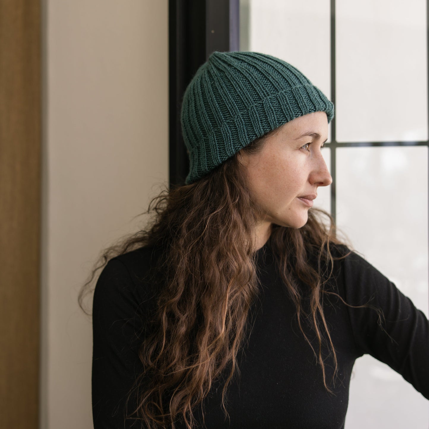 Woman wearing a green knit beanie and black long-sleeve shirt standing indoors.